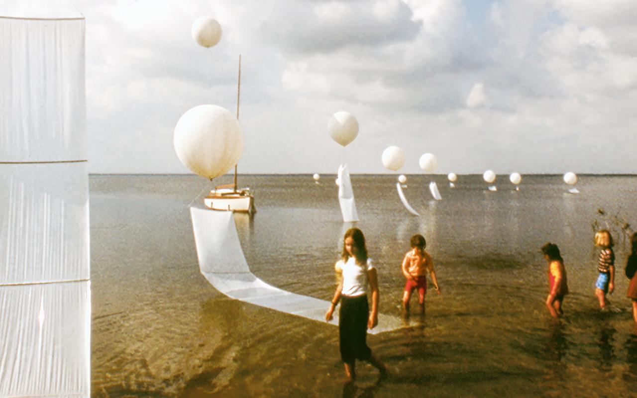 Zu sehen sind fünf Kinder, die durch Wasser laufen. Auf der linken Seite sind weiße Luftballons zu sehen.