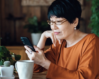 Lady wearing cooper color shirt and glasses is sitting at dinning table having coffee looking at her cell phone.