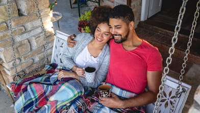 A couple on a porch swing under blankets holding mugs 