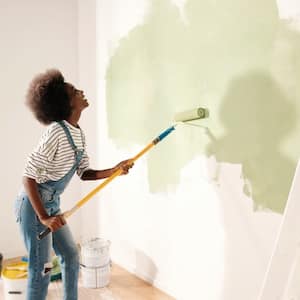 a woman in overalls painting a white wall with green paint and smiling