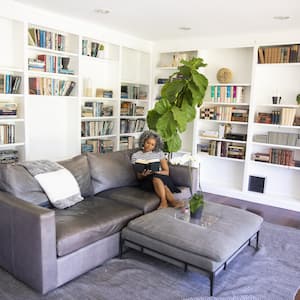 Black woman with curly white hair reading a book on a gray leather couch in her home library