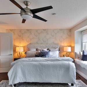 Ceiling fan in bedroom with gray paisley accent wall, gray bedding, and window seat with fur throw and pillows 