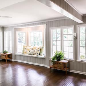 Large and open living room leading into sunroom with windows on two sides and lots of natural light flowing in. There is a window seat on one side and a leather couch and plant on the other.