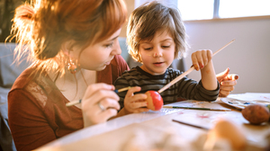 Little boy sitting by a table, on his mothers lap, painting an easter egg red.