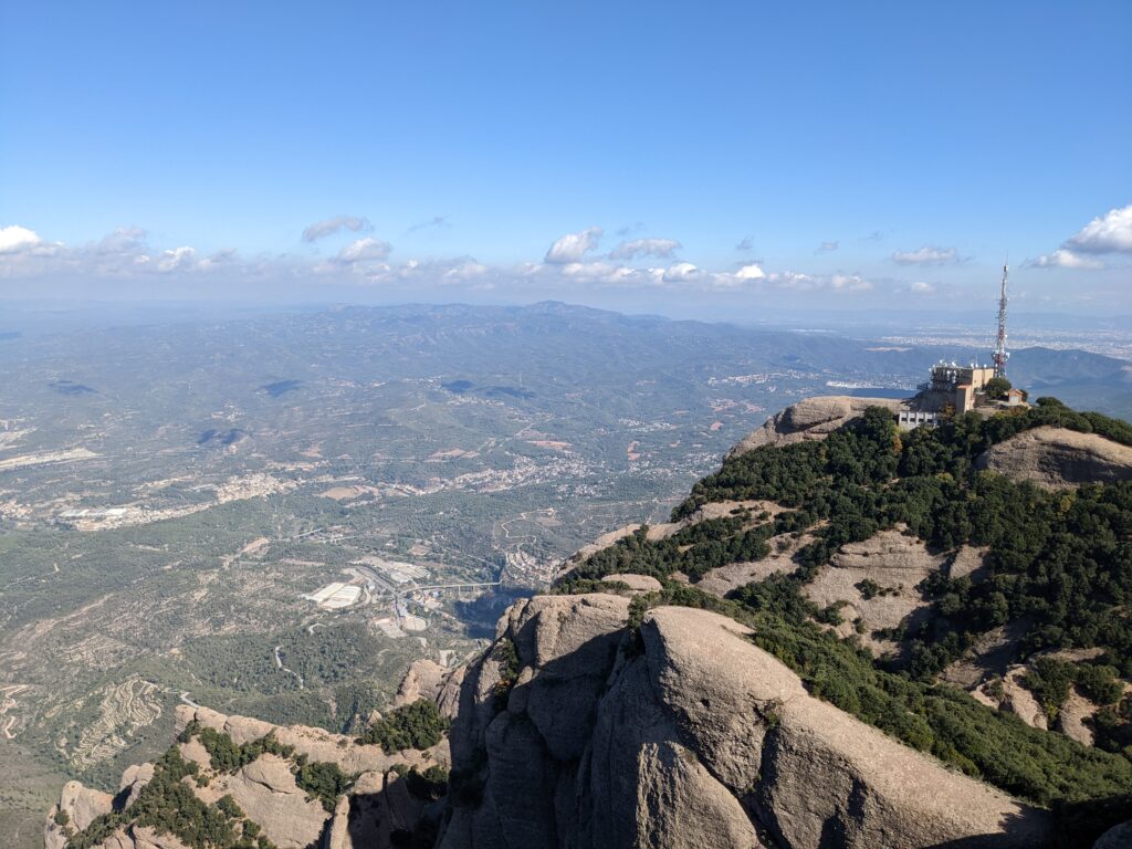 Picture from the peak of a mountain: rocky mountain top with bushes occupying the lower right quadrant of the image, a radio tower on the right, east-Ibrian landscape as far as the eye can see, with blue haze covering mountains far in the distance to the horizon, blue skies, clouds in the distance.