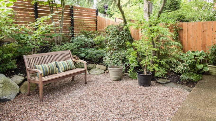 a light wood privacy fence surrounds a private yard with plants, bushes, a bench, and gravel