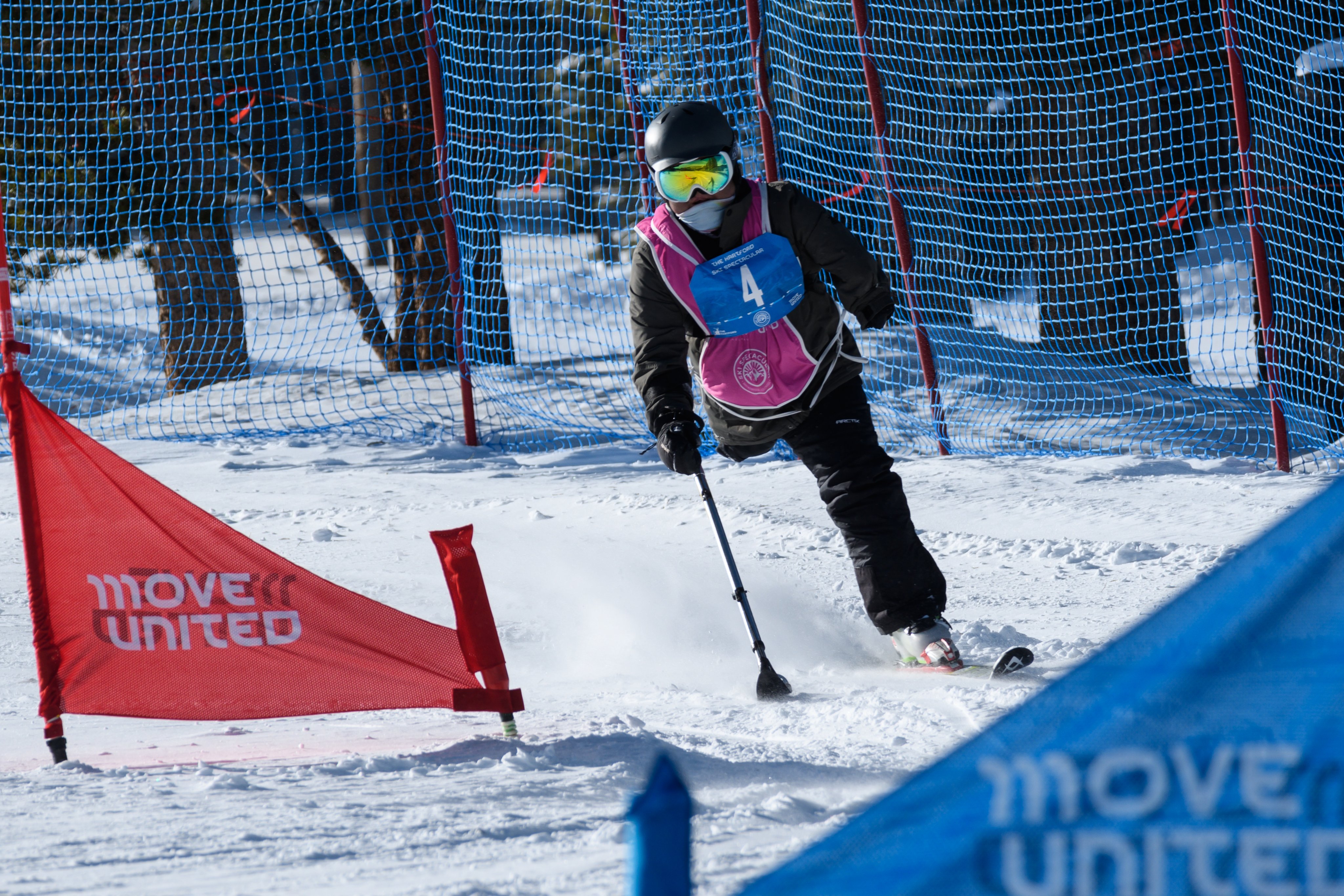 Individual with right leg amputation skiing down a mountain around flags that read Move United