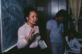 A female teacher stands in front of a blank blackboard while holding envelopes and gesturing with her hand. Two young men look at a book off to the side.