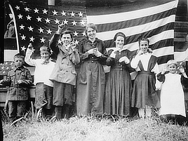 A group of seven smiling boys and girls stand in front of the American flag.