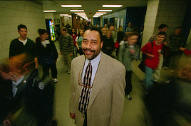 A man in a suit jacket and tie smiles while standing in a hallway with young people passing by around him.