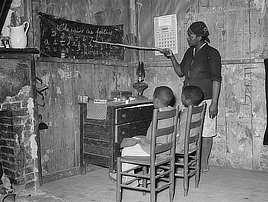 An African American woman  points at the number 7 in a home-made number and alphabet chart while two young children sit in chairs facing the chart.