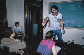 A male teacher in a t-shirt stands in front of a chalkboard illustrated with music notes and points towards adult students sitting in chairs.