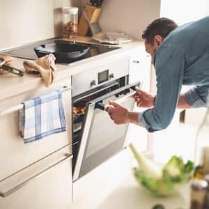 man opening oven to check on food inside in bright, cream colored kitchen strewn with various cooking tools