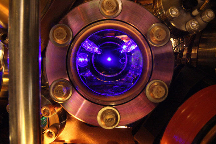 Window to the chamber of a scientific instrument. Inside at its center, a small ball of light illuminates the interior of the chamber in a purple glow.