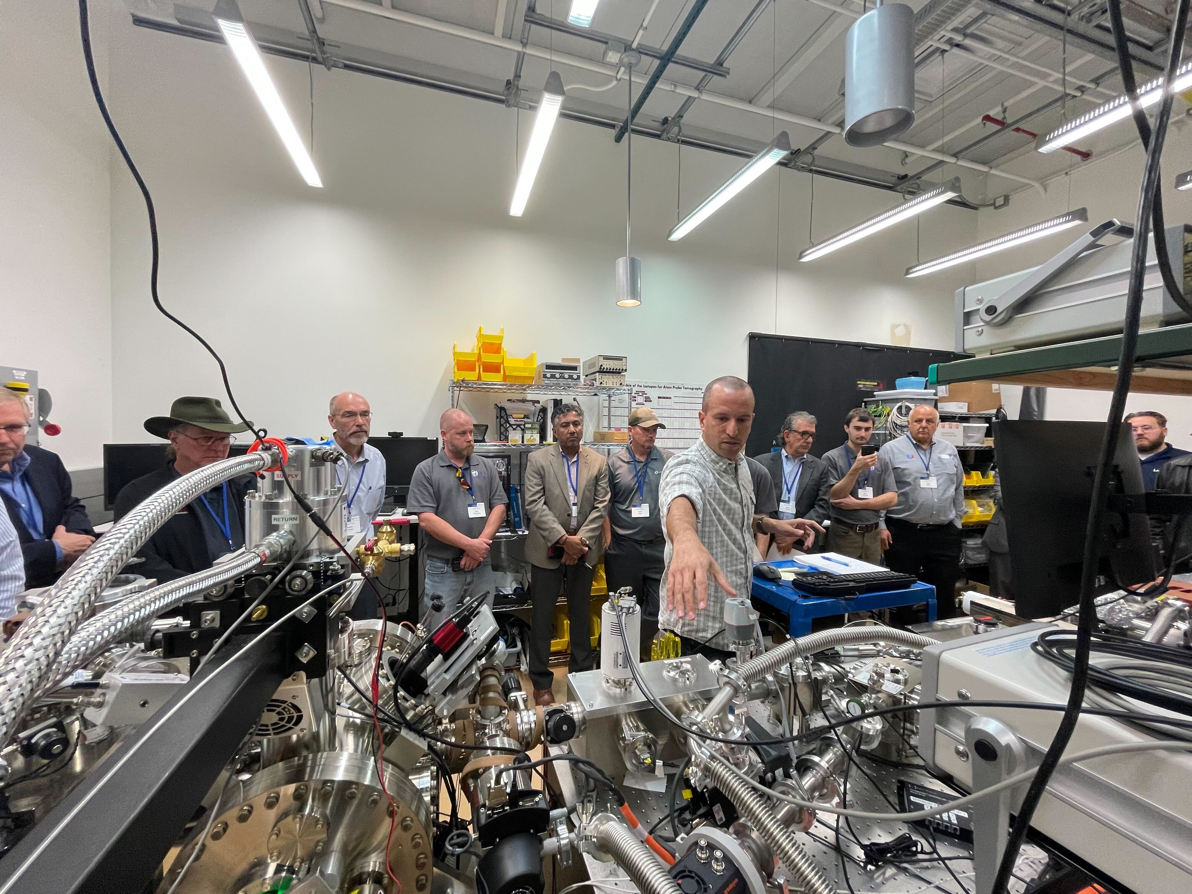 Scientist in a laboratory gestures toward multiple instruments. A group of people stand in the background, listening.