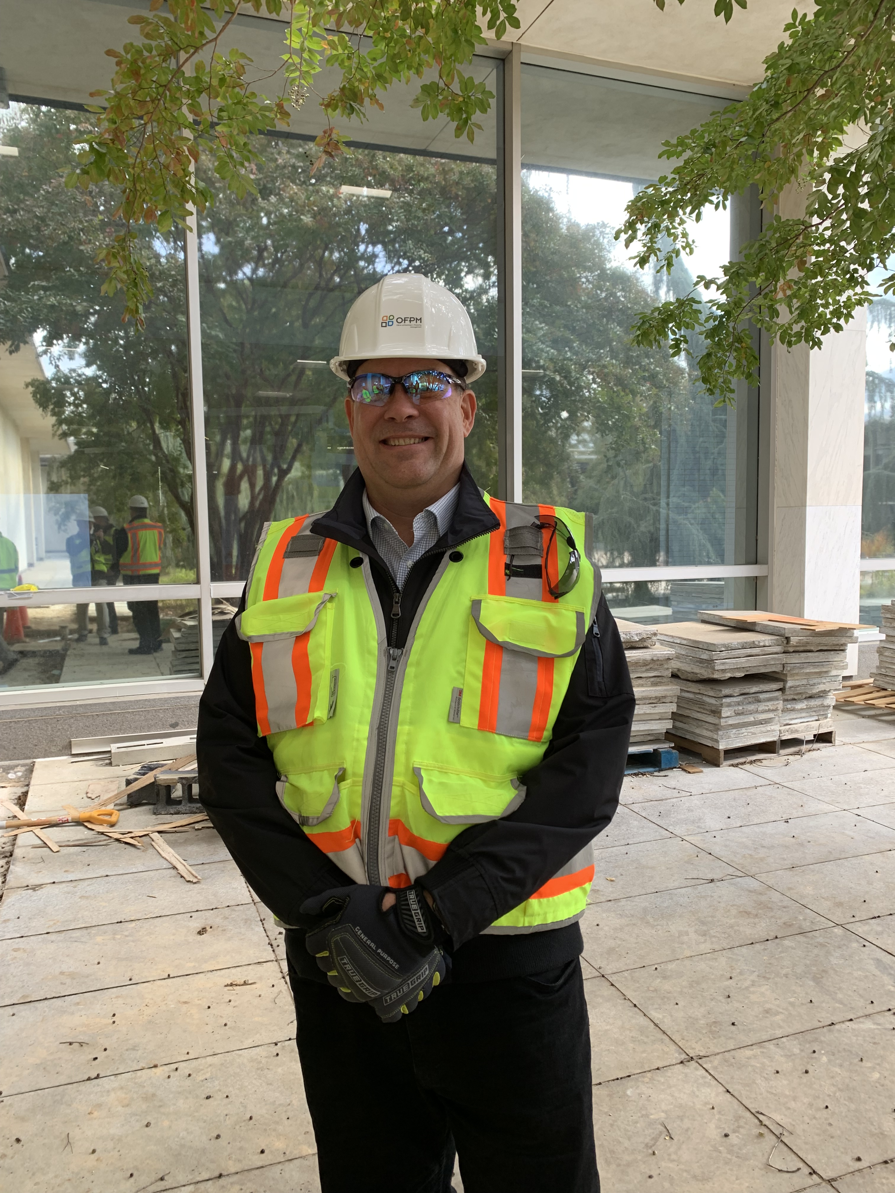 Man wearing a hard hat, safety glasses and a reflective yellow vest stands outside of a building, in front of a pile of construction supplies.