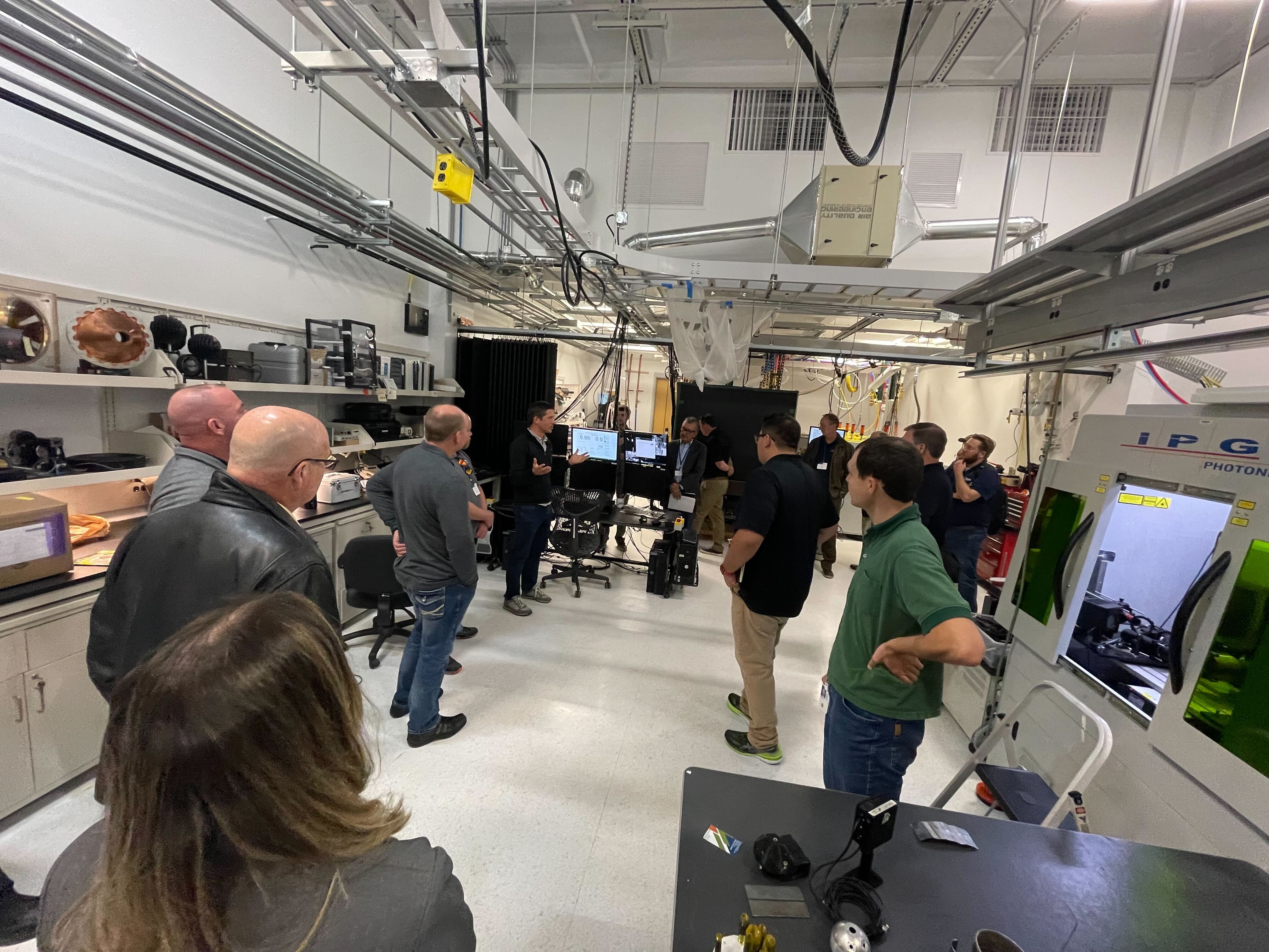 A group of people in a laboratory setting stand and listen to a person speaking.