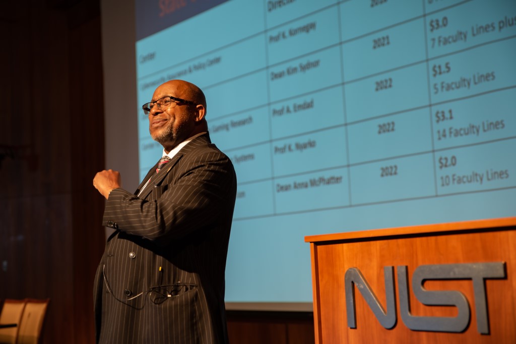 Willie May stands next to a podium that reads "NIST," speaking in front of a projector screen.