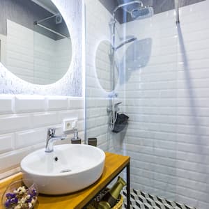 Modern small bathroom with white tile walls, glass rain shower, backlit round mirror, and vessel sink on narrow wood table
