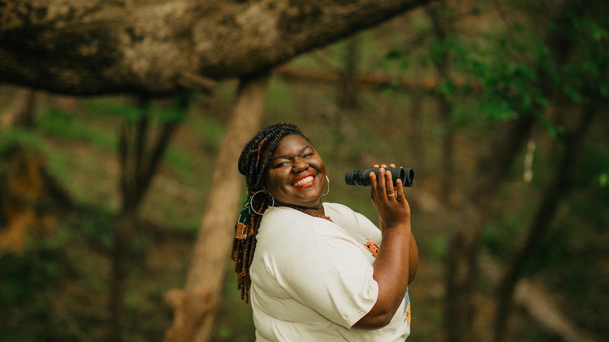 Bring Birds Back host Tenijah Hamilton smiling and holding binoculars in a wooded setting