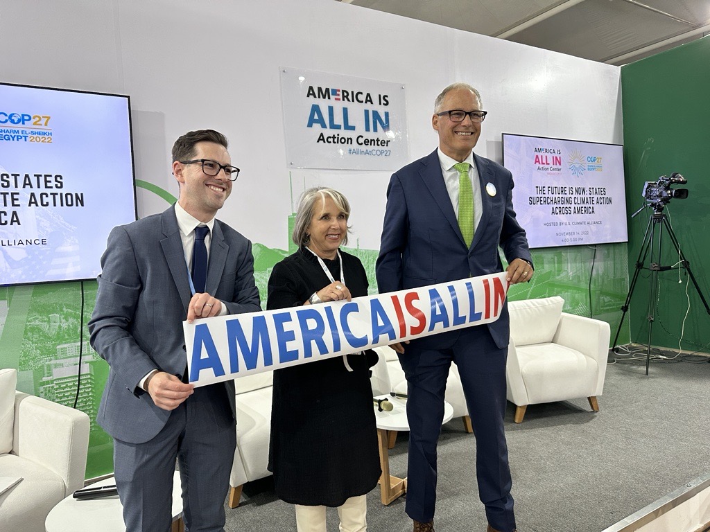 Two males with glasses and one female stand on a stage all holding a sign that reads 'America is all in.' Behind them are white chairs and two screens on a white and green wall.