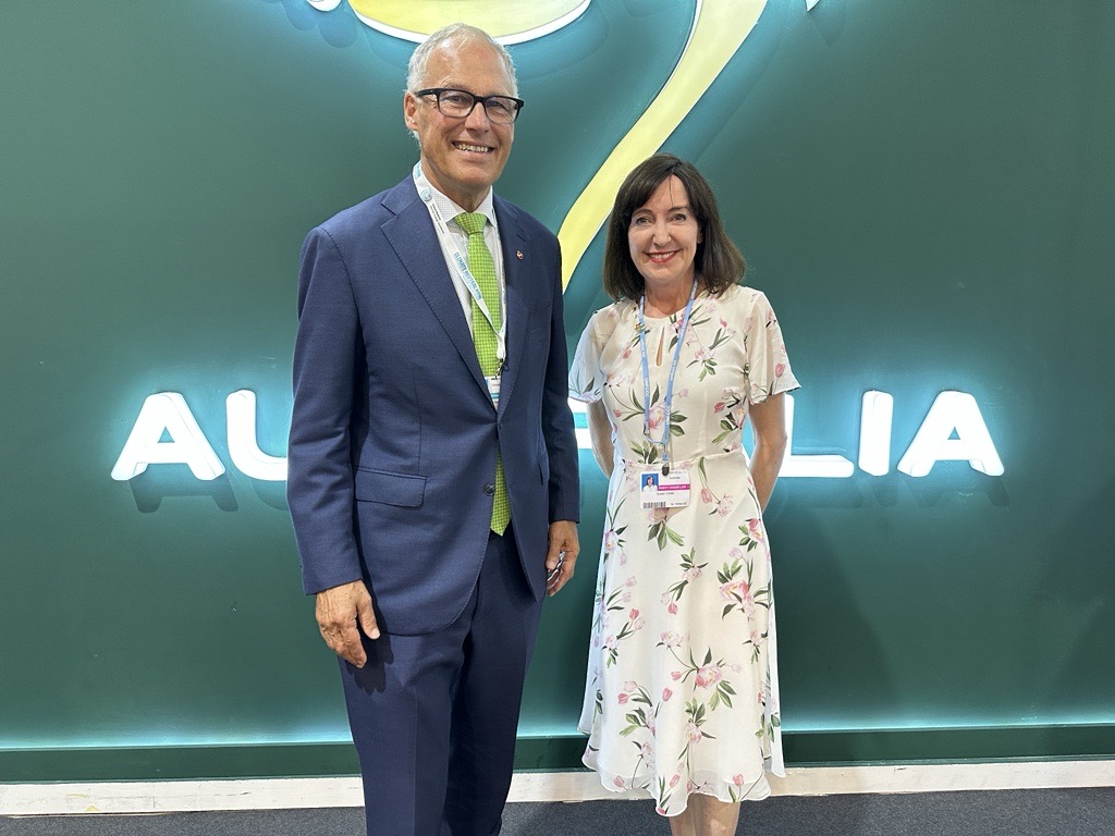 A man in a suit and a woman in a dress stand smiling for the camera in front of a large green wall with bright letters that read 'Australia.'