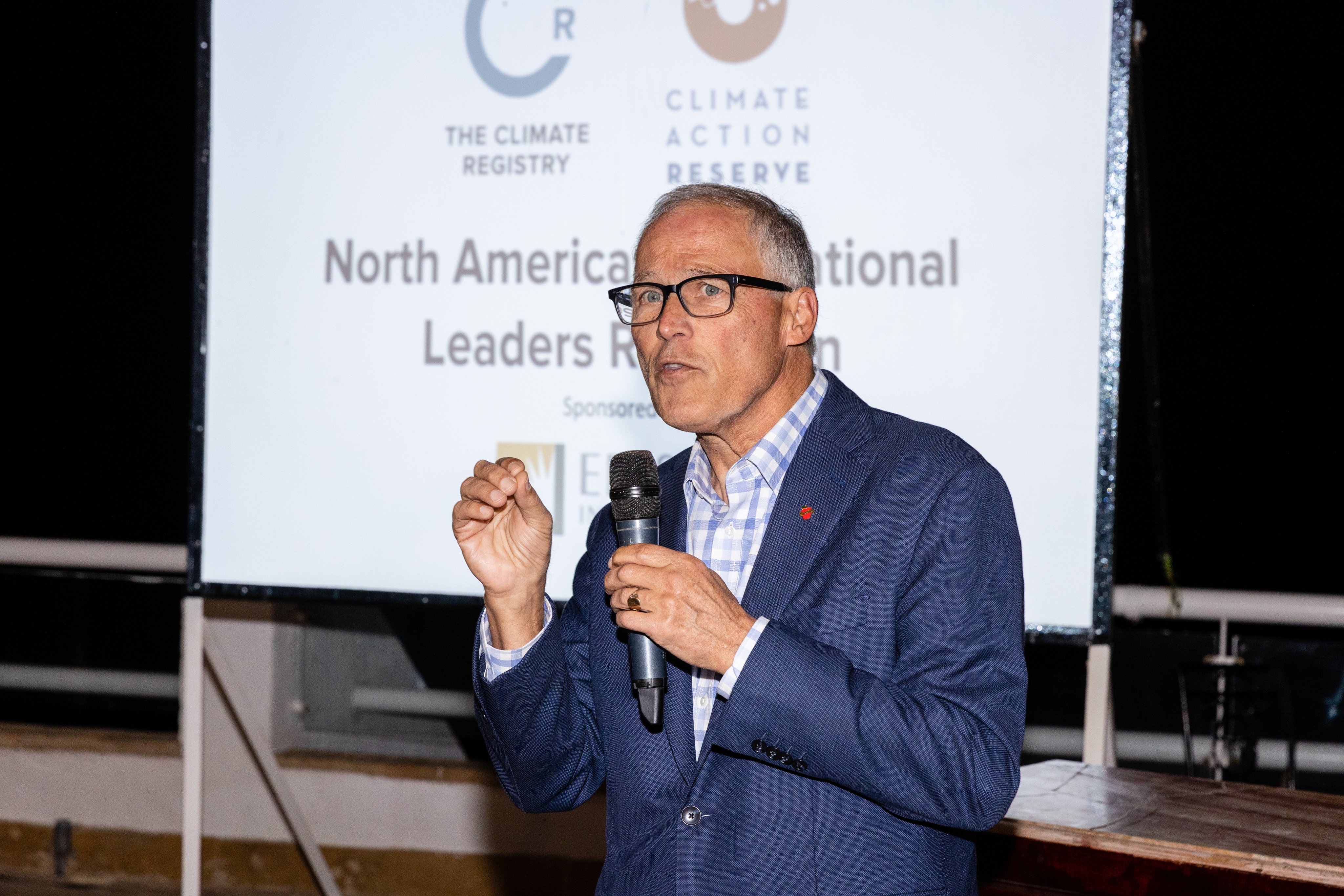 An older man with white hair and glasses stands with a microphone in his left hand addressing a room. Behind him is a large white screen that reads, 'North American National Leaders Reception.'
