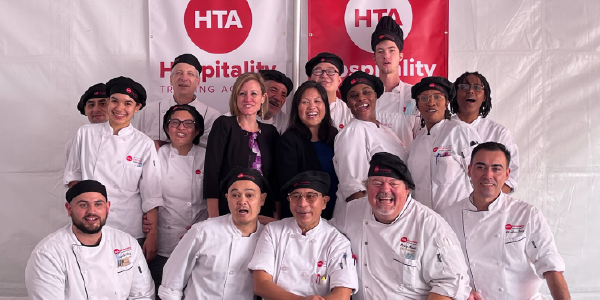 Julie Su smiles in the center of a group photo, surrounded by a diverse group of people in white chef coats. Banners behind them read: HTA, Hospitality Training Academy.