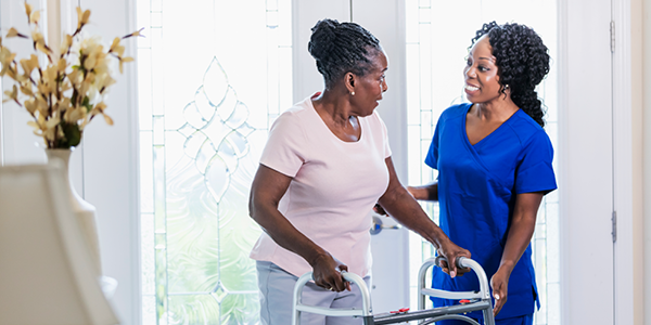 A woman in scrubs helps an older woman use a walker in the entrance of a house.