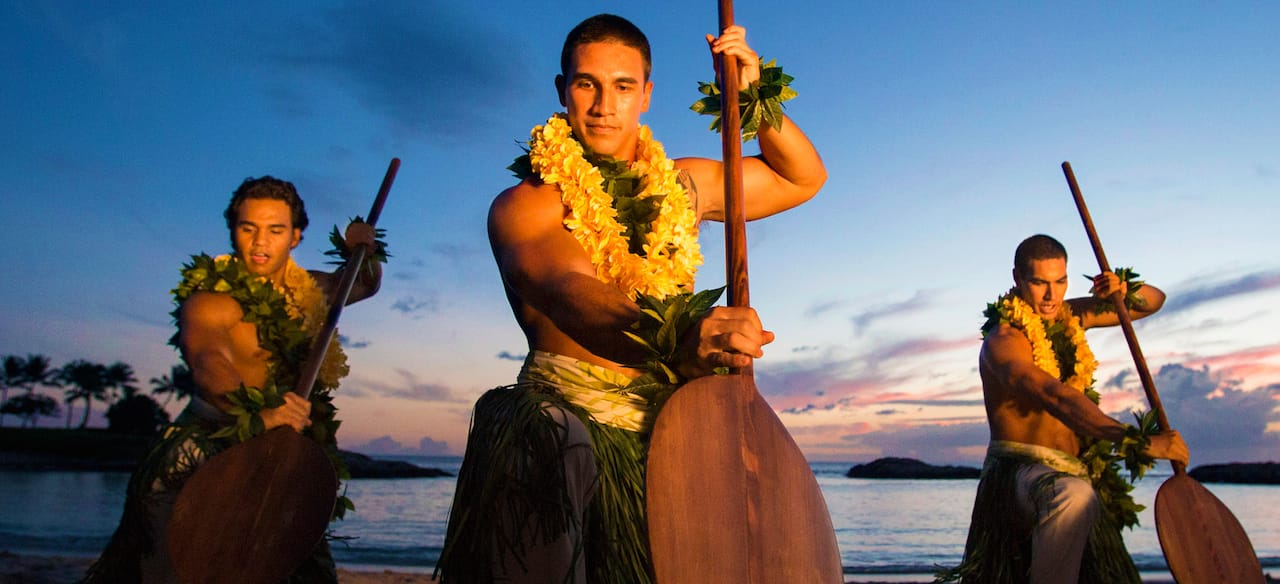 Three men wearing grass skirts, leis and maile leaves pose with large paddles on a beach at sunset