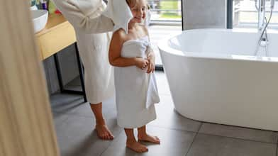 child in white towel gets hair dried by adult in bathroom with gray tiled floors and white bathtub