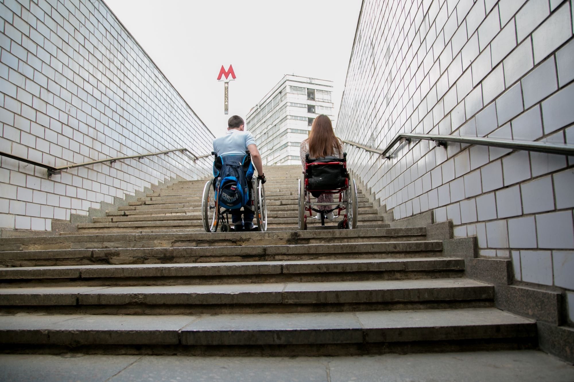 Two people in wheelchairs seen from the back trying to climb street stairs.