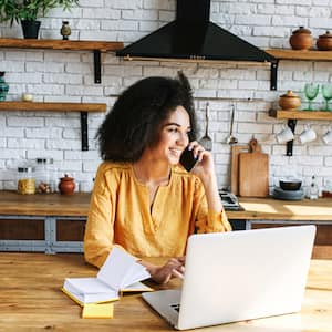 Black woman in her kitchen talking on phone at laptop