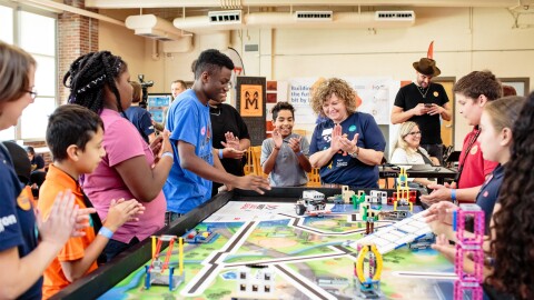 A group of young students and their teacher clap and smile as they stand around a table of their projects.