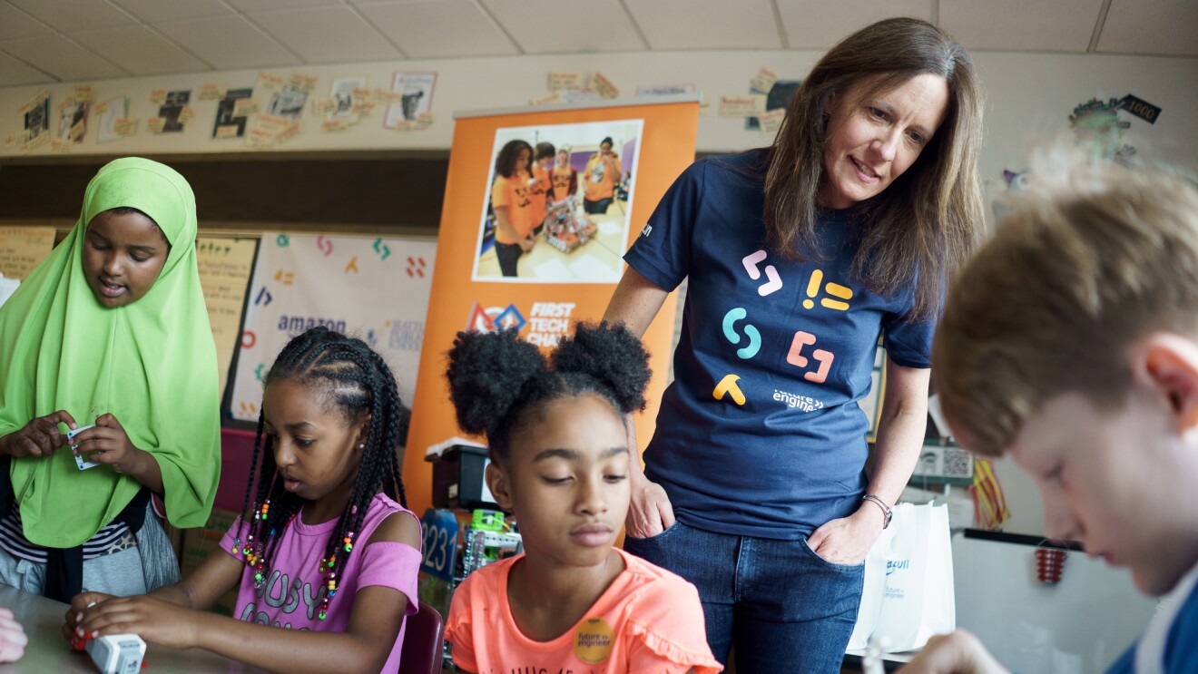 An Amazon employee wearing a Future Engineer t-shirt overlooks a group of students troubleshooting a device.