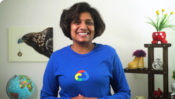 Woman in blue Google Cloud shirt smiling. In the background is a globe, a painting of a bird, and a plant.