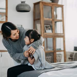 Young mother draping coat over her daughter’s shoulders inside their home