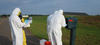 Four figures in coveralls stand outdoors around a mailbox, looking at a clipboard and other items. 