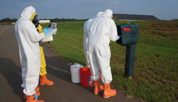 Four figures in coveralls stand outdoors around a mailbox, looking at a clipboard and other items. 