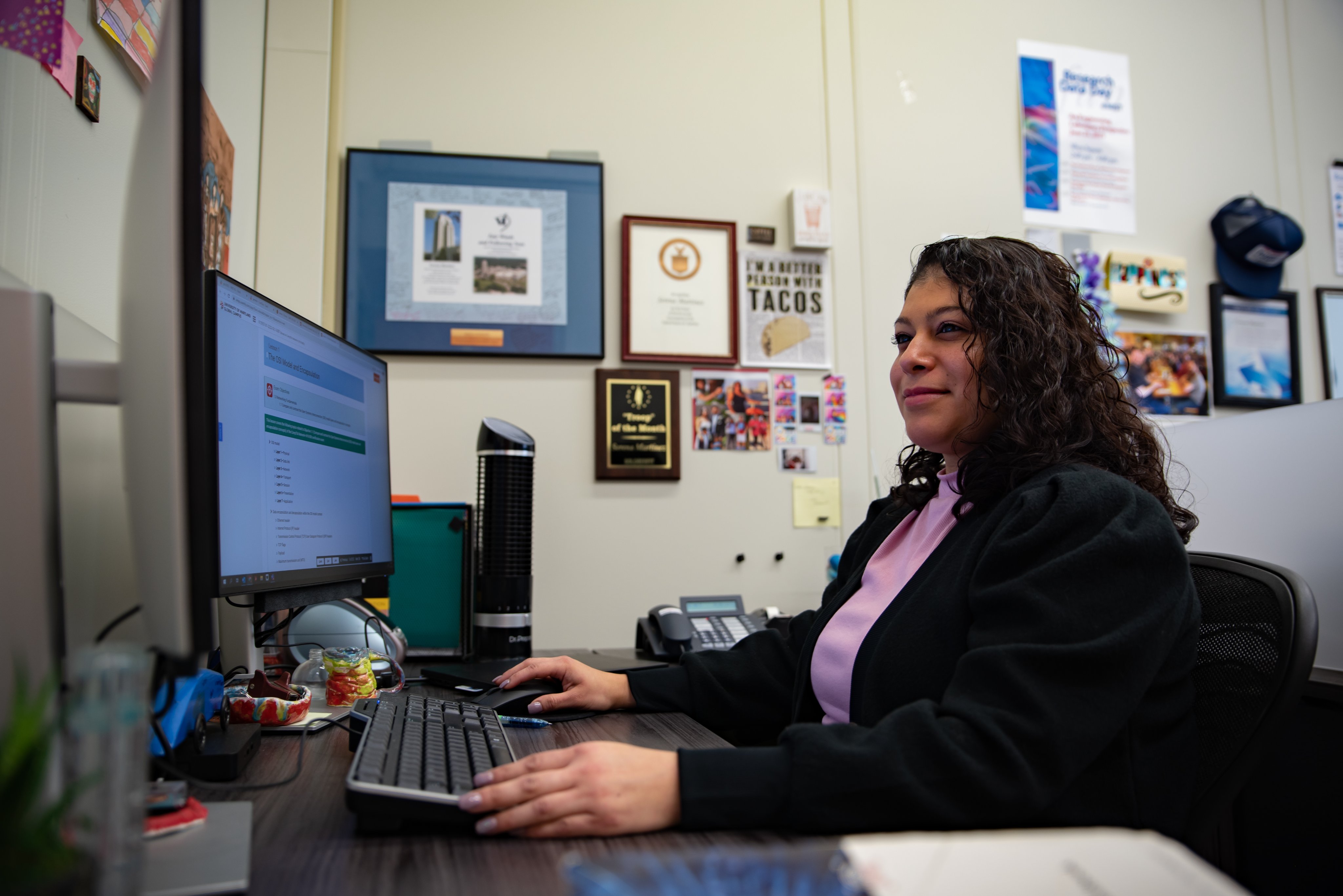 Woman sits at a desk in an office space and works at a computer station.