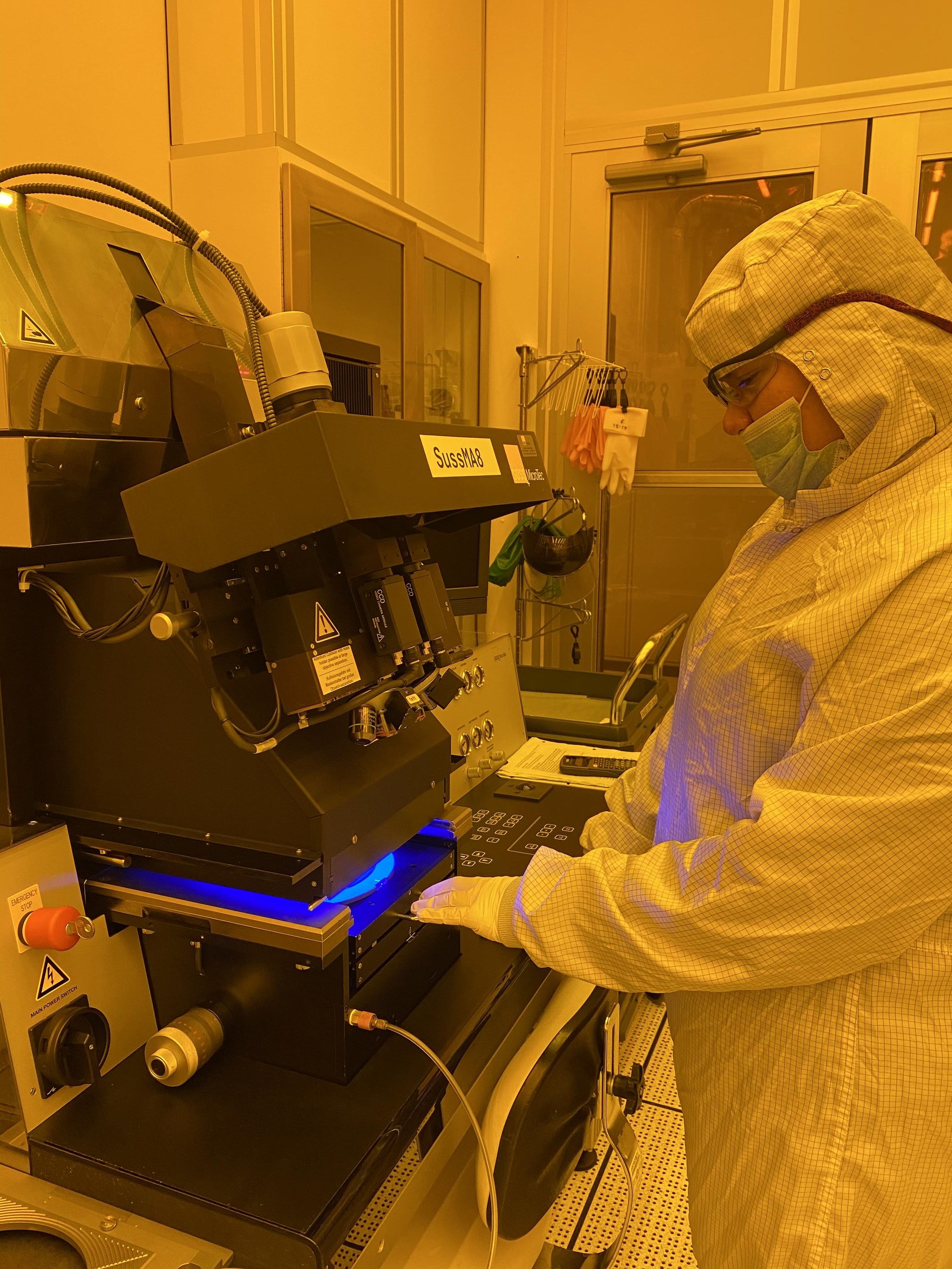 A person in white coveralls and safety glasses looks down at a machine she is using in a lab with orange lighting.