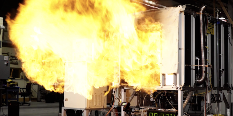 A fireball rushes out of the opening of a small metal compartment in a concrete room. 