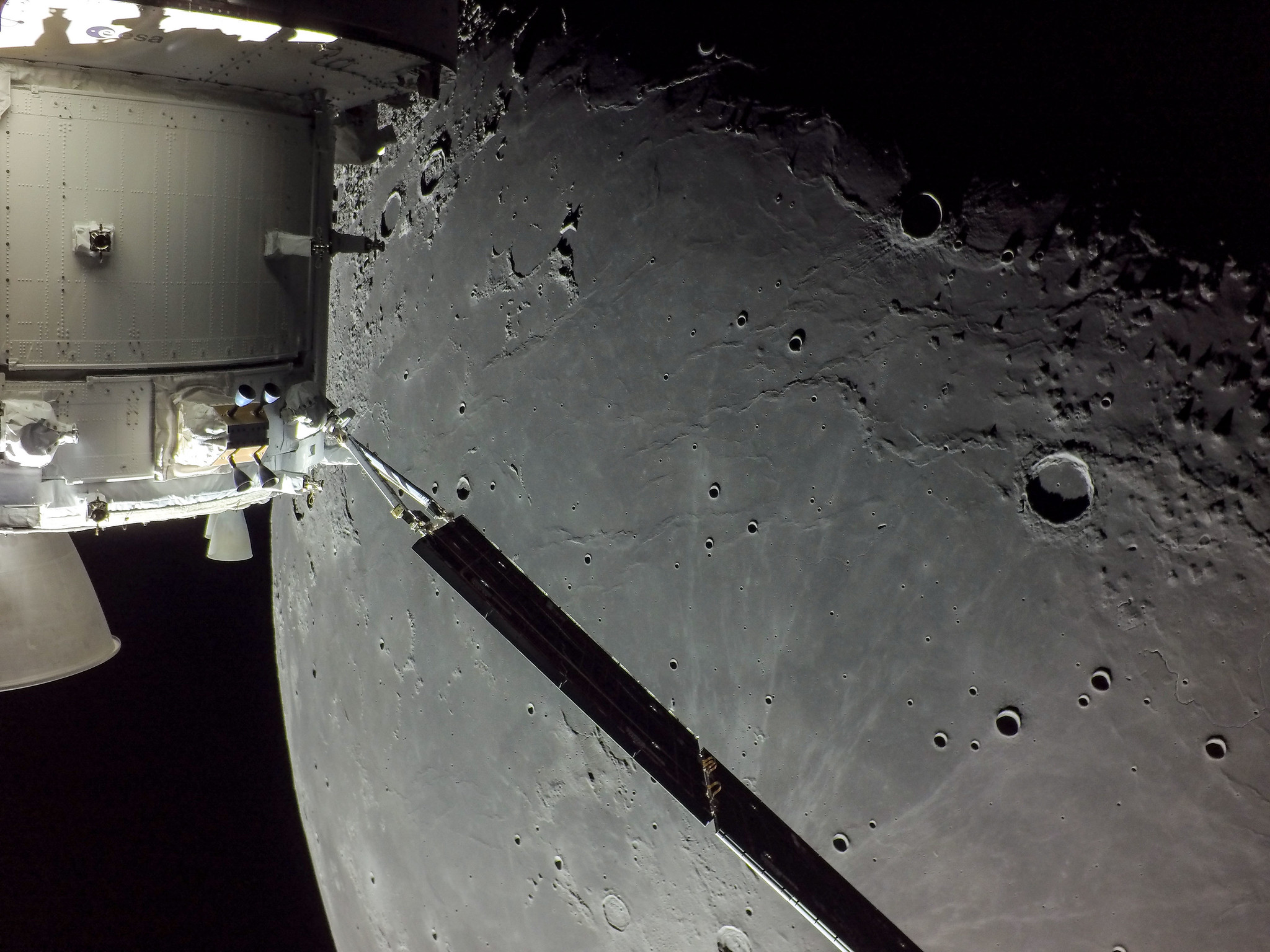 A portion of the Moon looms large just beyond the Orion spacecraft in this image taken on the 20th day of the Artemis I mission by a camera on the tip of one of Orion’s solar arrays. Craters are visible on the grey lunar surface. The blackness of space can be glimpsed behind the Orion spacecraft and the Moon. 