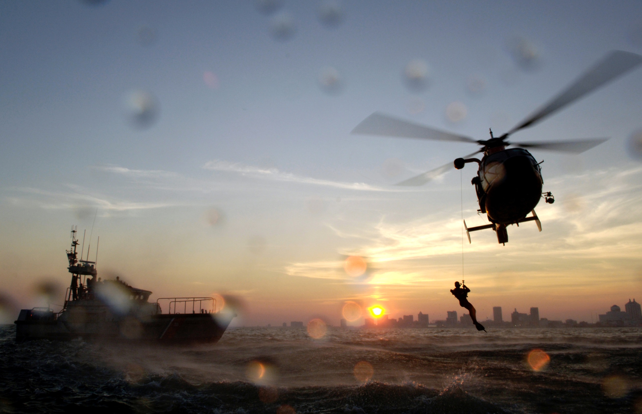 A helicopter flies low above the ocean during sunrise. A rescue diver is suspended from the helicopter, and nearby, a boat floats on the surface of the water. The skyline of a city can be seen in the far distance against a sky of blue and gold hues. Water droplets can be seen on the lens of the camera capturing the image. Credit: U.S. Coast Guard