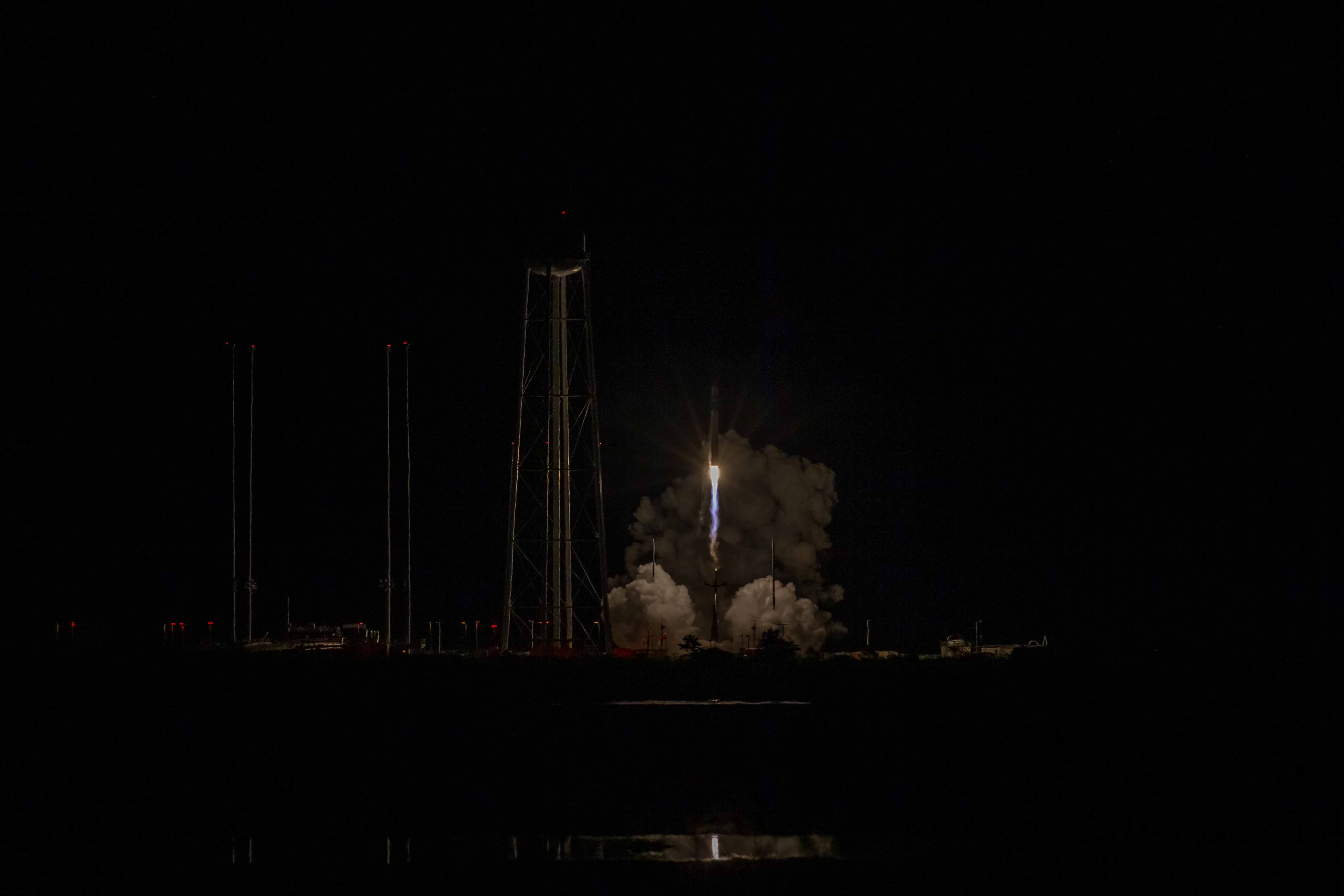 Rocket Lab's small black and white rocket launches off the pad in near pitch black. The rocket is just off the pad, surrounding by smoke on the bottom left, right, and just behind it. A water tower with spindly legs and four tall poles are to the left of the rockett, slightly lit up by the blow of the rocket. A small reflection of the rocket is seen toward the bottom of the image in water.