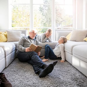 elderly man and two young kids read book in front of large windows between two white couches