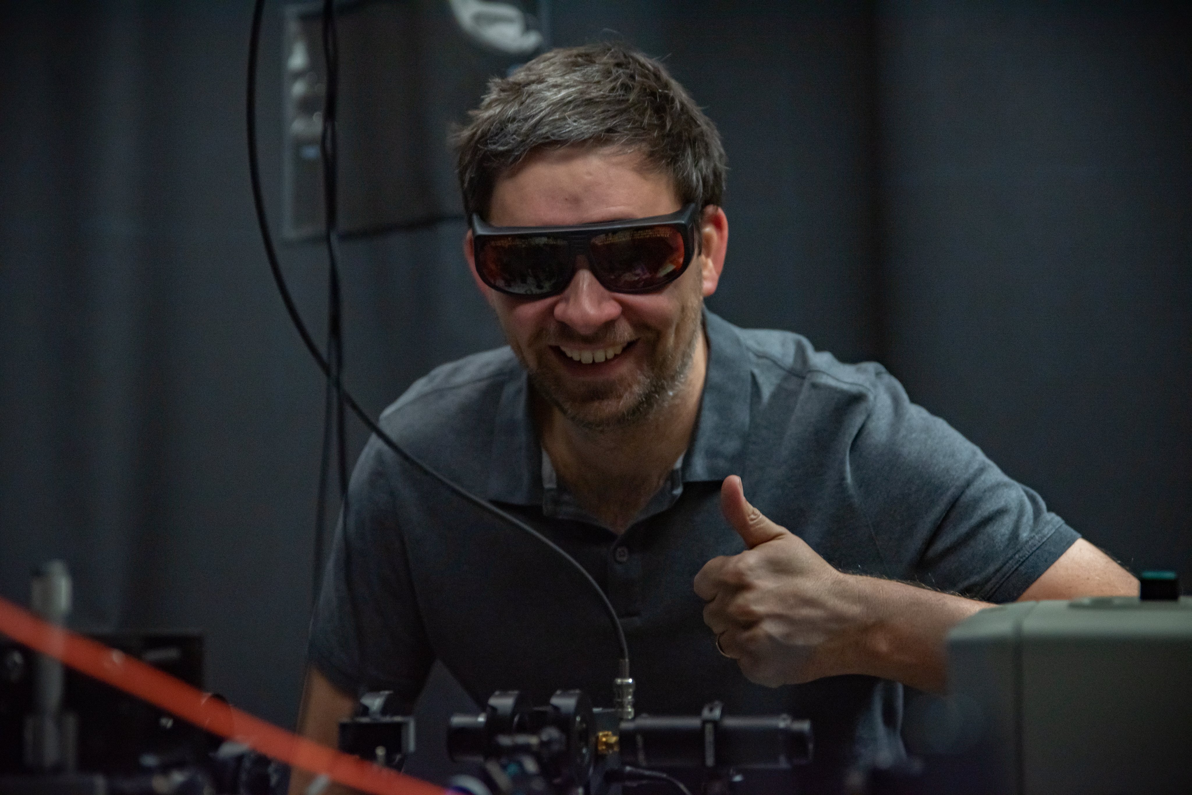 Man wearing safety glasses in a laboratory setting smiles and holds a thumbs up.