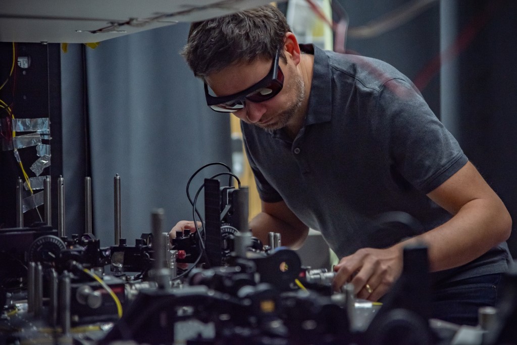 Man wearing safety glasses leans toward a tabletop optical laser setup.