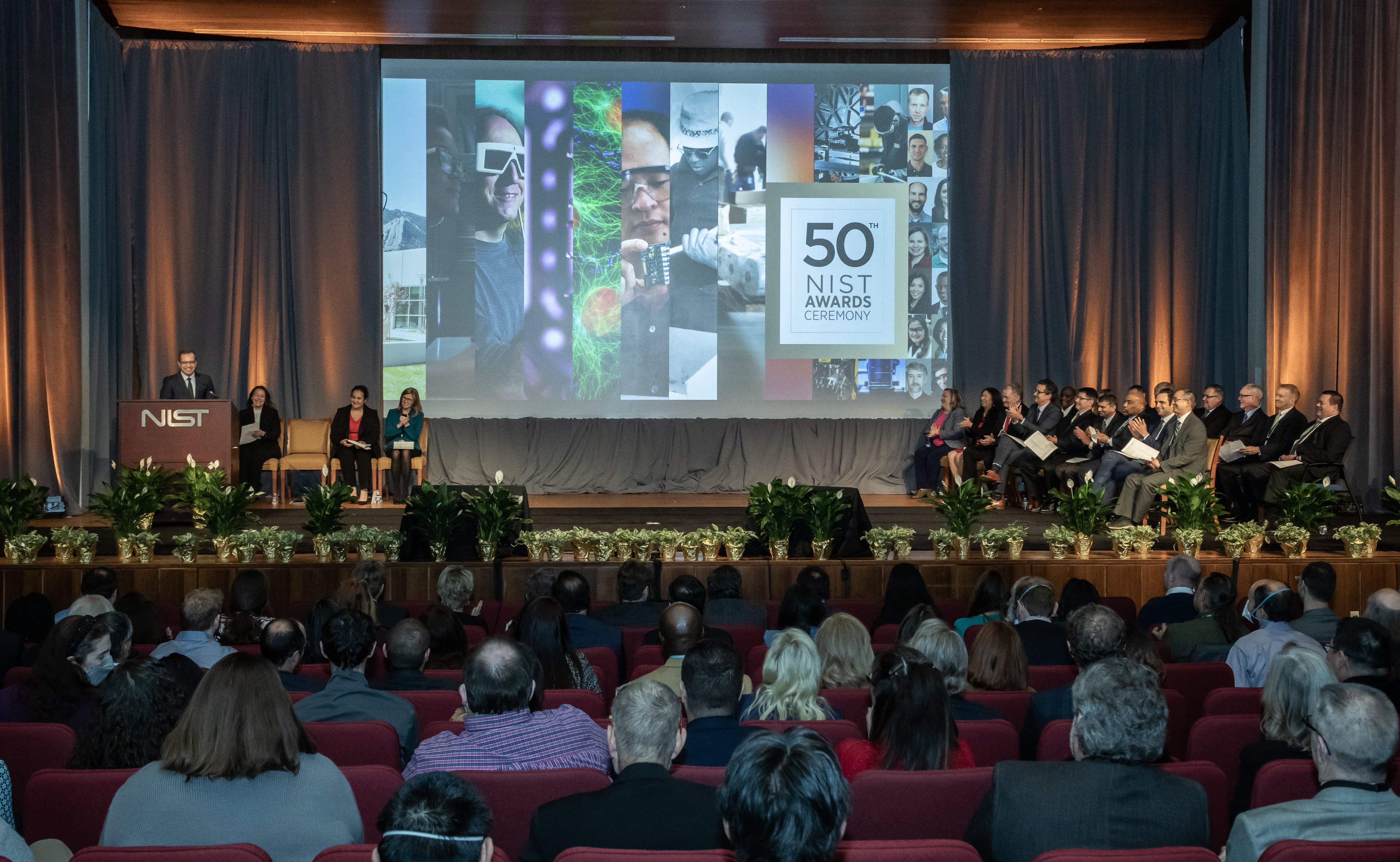 A full auditorium. A screen on stage shows a collection of photos alongside text that reads: "50th NIST Awards Ceremony."