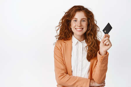 Smiling redhead saleswoman shows credit card and looks confident, pay contactless, stands against white background in suit